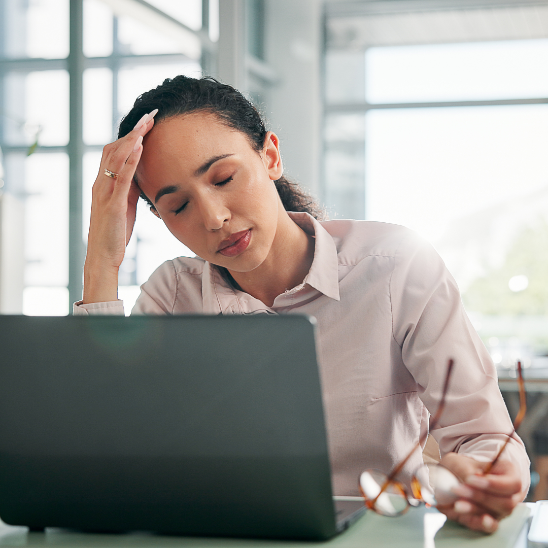 Donna seduta alla scrivania davanti al computer, con gli occhi chiusi e una mano sulla fronte, mentre manifesta stanchezza e stress da rientro al lavoro.