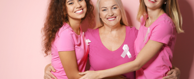 Tre donne di diversa età indossano magliette rosa con il fiocco simbolo della prevenzione contro il cancro al senso e si abbracciano sorridendo, su uno sfondo rosa.