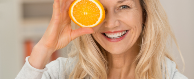 Donna sorridente che tiene in mano un'arancia e una mezza arancia davanti a un occhio, a rappresentare l'importanza di un'alimentazione ricca di nutrienti per sostenere il sistema immunitario