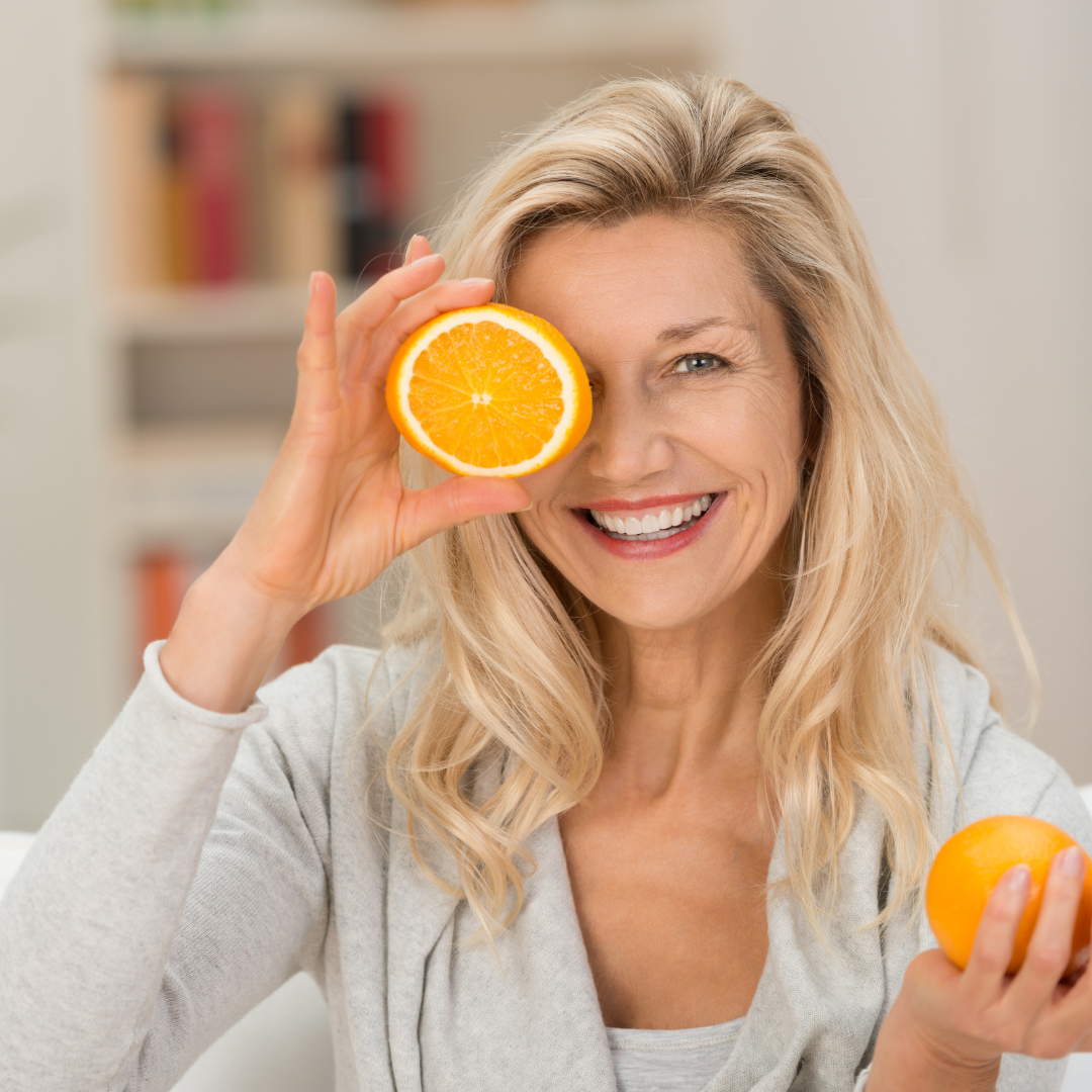 Donna sorridente che tiene in mano un'arancia e una mezza arancia davanti a un occhio, a rappresentare l'importanza di un'alimentazione ricca di nutrienti per sostenere il sistema immunitario